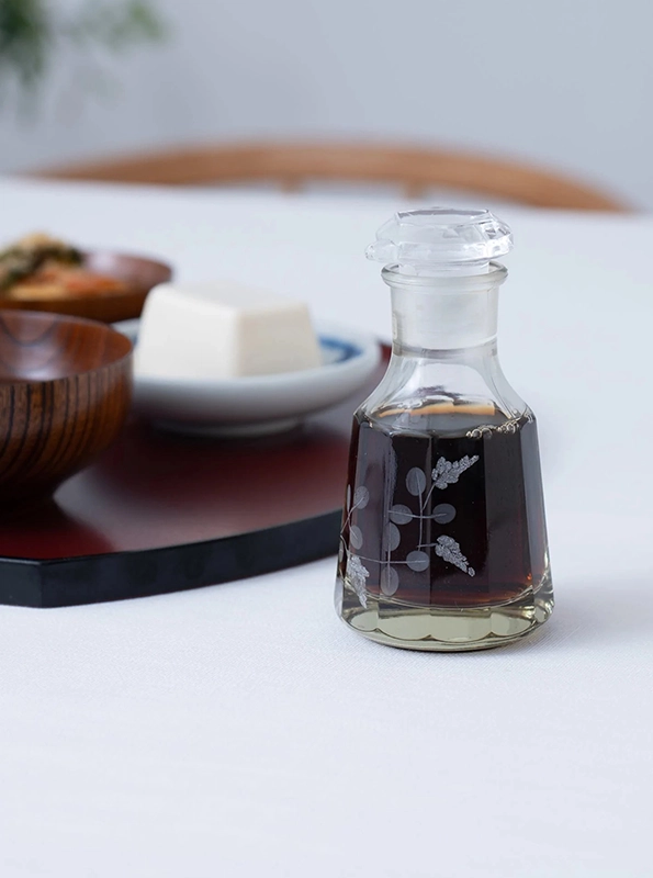 Μπουκάλι με σάλτσα σόγιας σε γυάλινη καράφα με διακοσμητικά σχέδια, τοποθετημένο σε τραπέζι, με πιάτα ιαπωνικής κουζίνας στο φόντο.