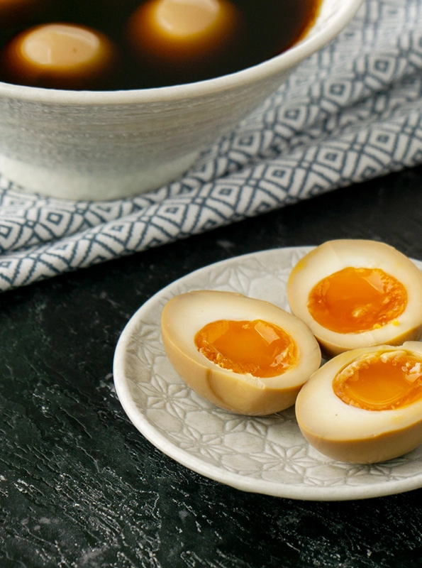 Μαριναρισμένα αυγά ράμεν (ajitsuke tamago) κομμένα στη μέση, με μαλακό, ρευστό κρόκο, σερβιρισμένα σε πιάτο, με μπολ μαρινάδας στο φόντο.