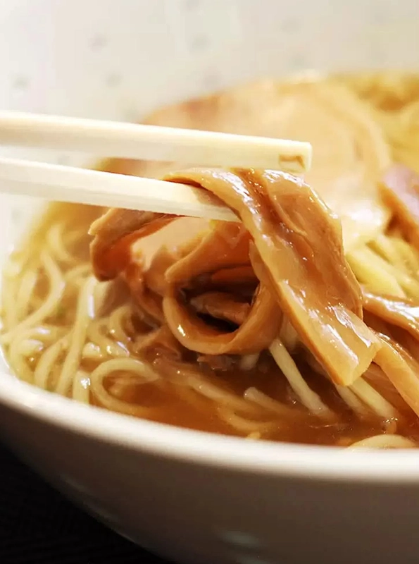 Κοντινό πλάνο από chopsticks που κρατούν menma, ζυμωμένους βλαστούς μπαμπού, πάνω από μπολ ράμεν με ζωμό και νουντλς.