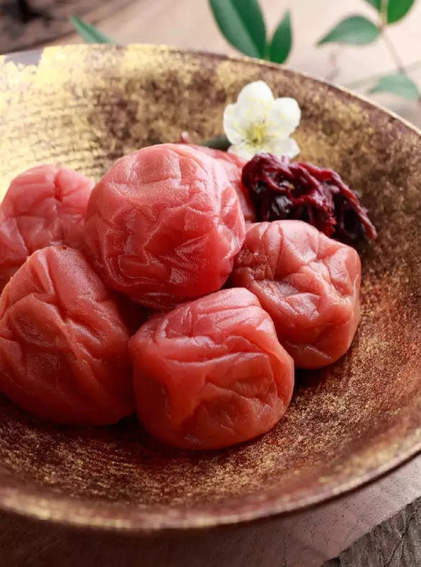 Κοντινό πλάνο από ιαπωνικά umeboshi – δαμάσκηνα τουρσί με έντονο κόκκινο χρώμα και ζαρωμένη υφή, σερβιρισμένα σε διακοσμητικό μπολ με λουλούδι και φύλλα shiso.