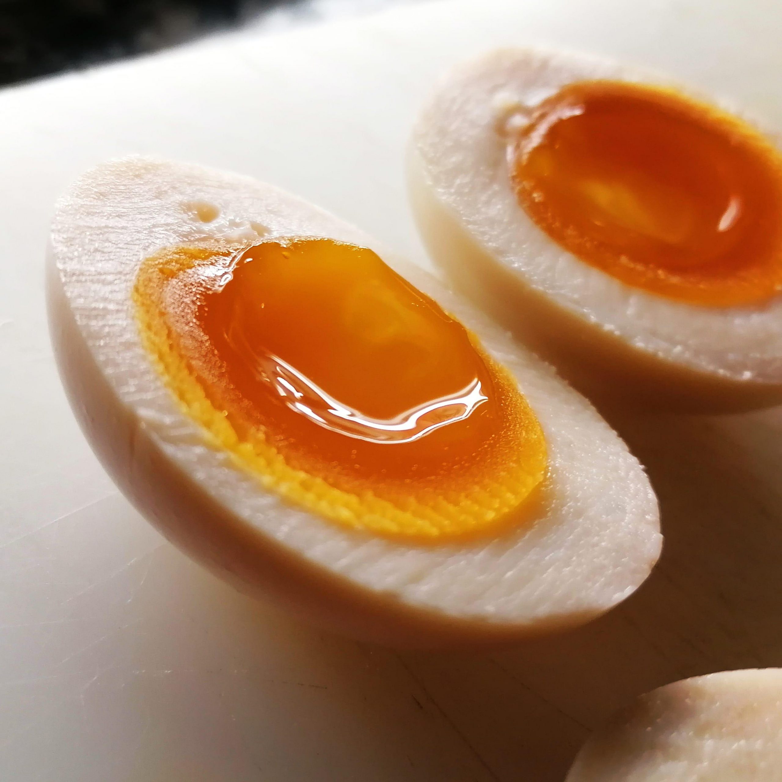 Κοντινό πλάνο δύο μισών αυγών ράμεν (ajitsuke tamago) με μελάτο, λαμπερό κρόκο πάνω σε λευκή επιφάνεια.
