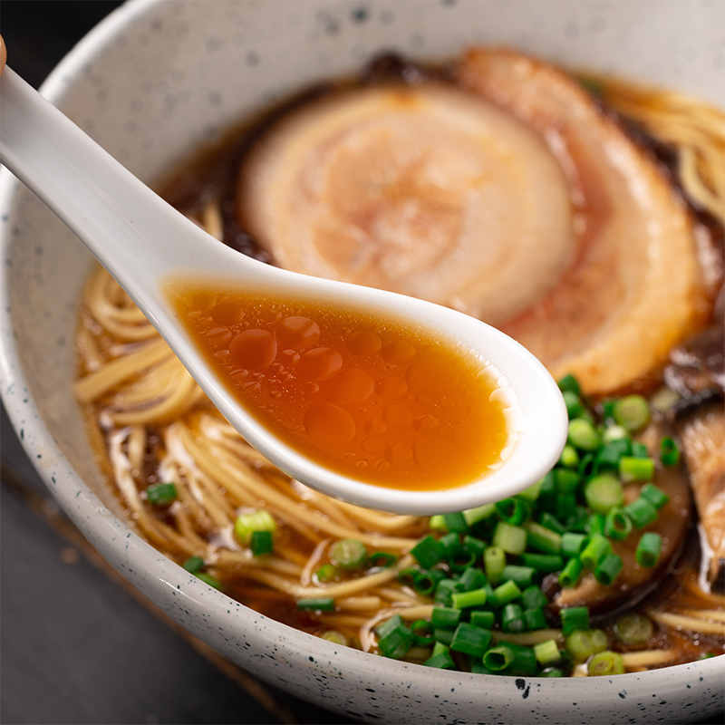 Κουτάλι με ζωμό shoyu ramen πάνω από μπολ με noodles, chashu χοιρινό και φρέσκα κρεμμυδάκια.