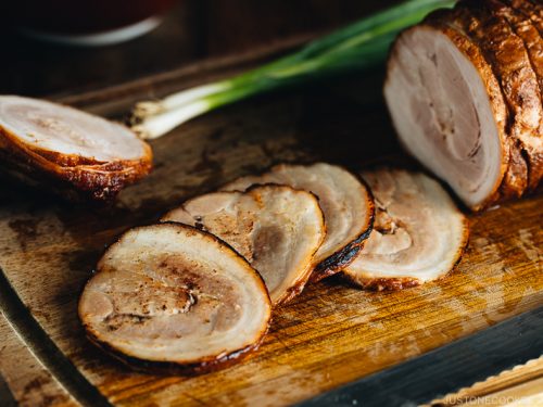 Φέτες ζουμερού chashu από χοιρινή κοιλιά, σιγομαγειρεμένες και καραμελωμένες, σε ξύλινη επιφάνεια κοπής με πράσινο κρεμμύδι στο φόντο.