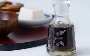 Μπουκάλι με σάλτσα σόγιας σε γυάλινη καράφα με διακοσμητικά σχέδια, τοποθετημένο σε τραπέζι, με πιάτα ιαπωνικής κουζίνας στο φόντο.