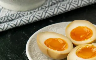 Μαριναρισμένα αυγά ράμεν (ajitsuke tamago) κομμένα στη μέση, με μαλακό, ρευστό κρόκο, σερβιρισμένα σε πιάτο, με μπολ μαρινάδας στο φόντο.