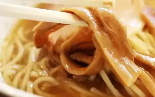 Κοντινό πλάνο από chopsticks που κρατούν menma, ζυμωμένους βλαστούς μπαμπού, πάνω από μπολ ράμεν με ζωμό και νουντλς.