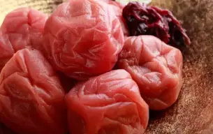 Κοντινό πλάνο από ιαπωνικά umeboshi – δαμάσκηνα τουρσί με έντονο κόκκινο χρώμα και ζαρωμένη υφή, σερβιρισμένα σε διακοσμητικό μπολ με λουλούδι και φύλλα shiso.