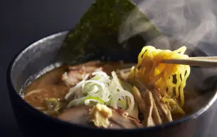 Αχνιστό μπολ ramen με κιτρινωπά noodles, φέτες χοιρινού chashu, φρέσκο κρεμμυδάκι, μπαμπού και φύκι nori, σε πλούσιο καφέ ζωμό.
