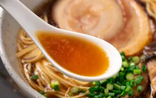 Κουτάλι με ζωμό shoyu ramen πάνω από μπολ με noodles, chashu χοιρινό και φρέσκα κρεμμυδάκια.