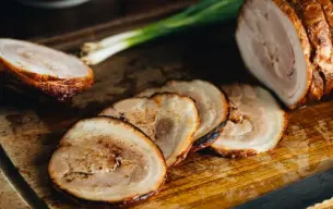 Φέτες ζουμερού chashu από χοιρινή κοιλιά, σιγομαγειρεμένες και καραμελωμένες, σε ξύλινη επιφάνεια κοπής με πράσινο κρεμμύδι στο φόντο.